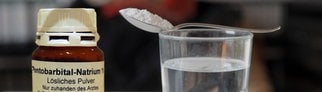 Eine Flasche Natrium-Pentobarbital, daneben ein Wasserglas mit einem Löffel, auf dem Pulver gehäuft ist