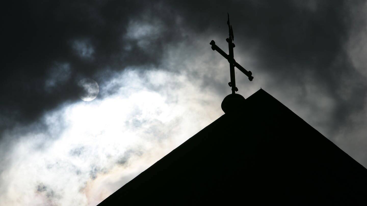 Im Gegenlicht und vor wolkenverhangenem Himmel ist die Kirchturmspitze des Doms in Osnarbrück mit Kreuz zu sehen.