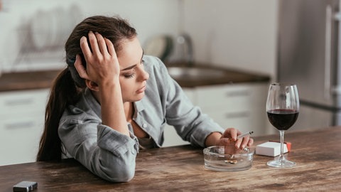 Junge Frau sitzt gestresst im Wohnzimmer, sie raucht eine Zigarette und hat ein Glas Wein auf dem Tisch stehen (Foto: Adobe Stock / LIGHTFIELD STUDIOS)