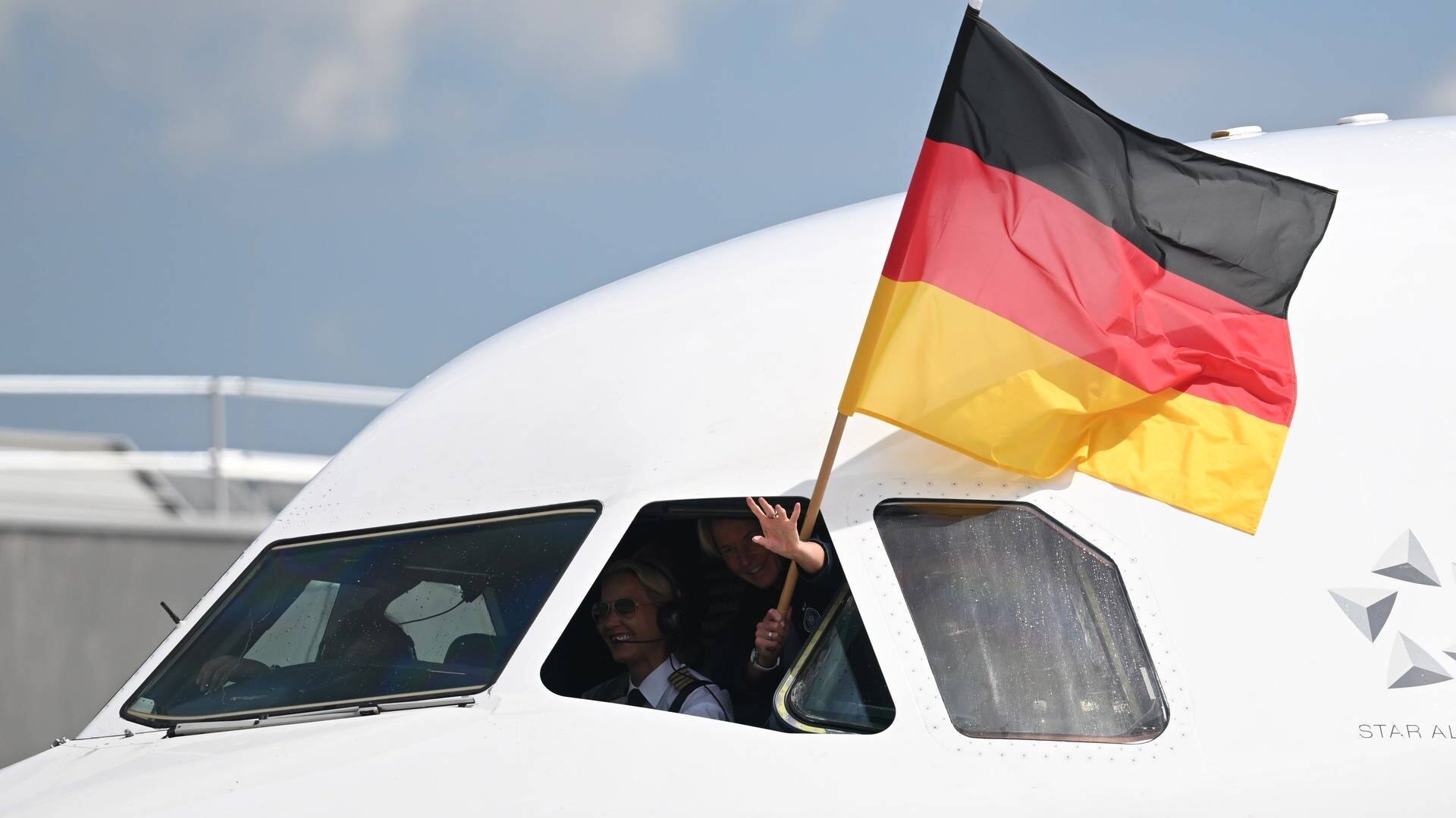 Mit einer Deutschlandflagge winkt Bundestrainerin Martina Voss-Tecklenburg neben der Pilotin aus dem Cockpitfenster. (Foto: dpa Bildfunk, picture alliance/dpa | Arne Dedert)