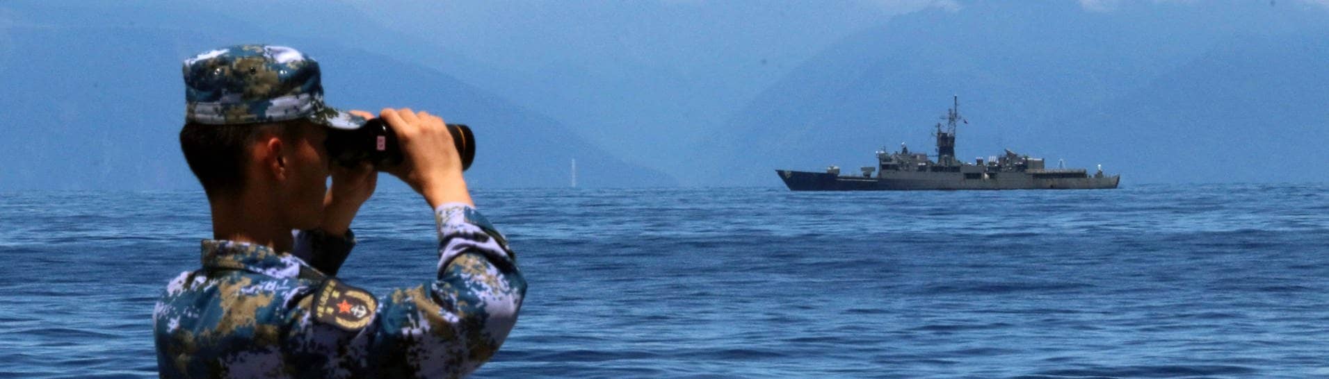 Ein Mitglied der chinesischen Volksbefreiungsarmee, das während einer Militärübung auf See durch ein Fernglas guckt, während im Hintergrund Taiwans Fregatte Lan Yang zu sehen ist. (Foto: dpa Bildfunk, picture alliance/dpa/Xinhua/AP | Lin Jian)