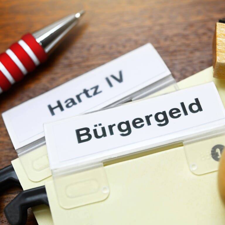 Auf einem Tisch liegen zwei Kärtchen auf denen „Bürgergeld“ und „Hartz IV“ steht. Daneben liegt ein Stempel und ein Kugelschreiber. (Foto: IMAGO, IMAGO / Christian Ohde)