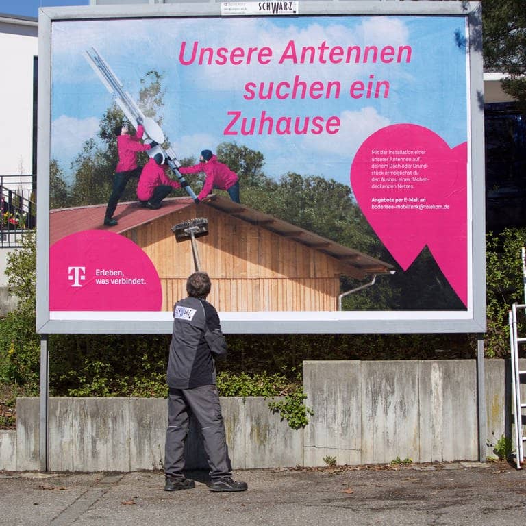 Ein Mann klebt ein großes Plakat fest, darauf ein Schriftzug, dass die Telekom neue Standorte für Antennen sucht (Foto: Deutsche Telekom)