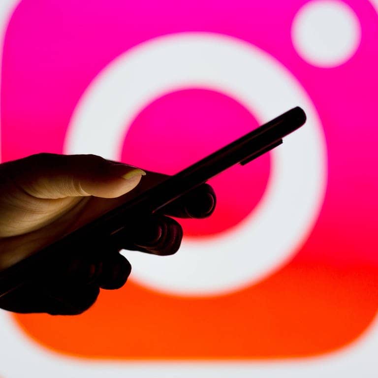 Silhouette einer Hand, die ein Smartphone hält, im Hintergrund das Instagram-Logo (Foto: IMAGO, IMAGO/Zoonar)