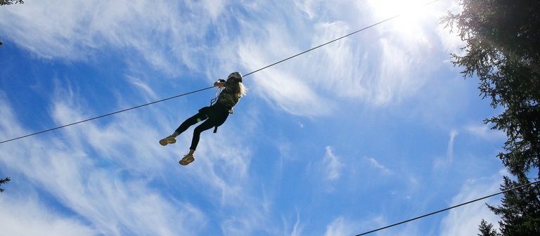 Eine junge Frau gleitet mit einer Seilrutsche vor blauem Himmel (Foto: IMAGO, imago images/Geisser)