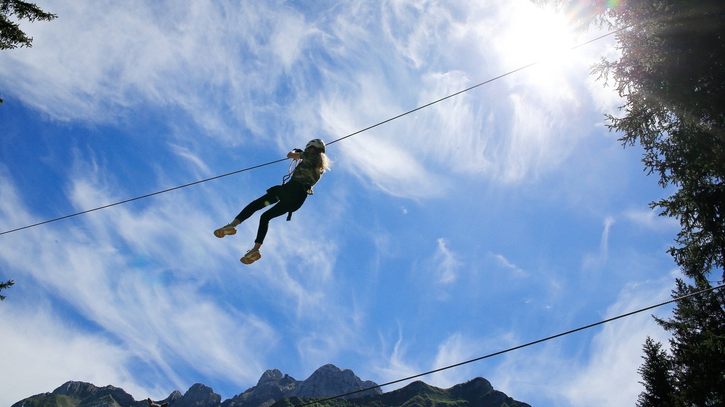 Eine junge Frau gleitet mit einer Seilrutsche vor blauem Himmel (Foto: IMAGO, imago images/Geisser)