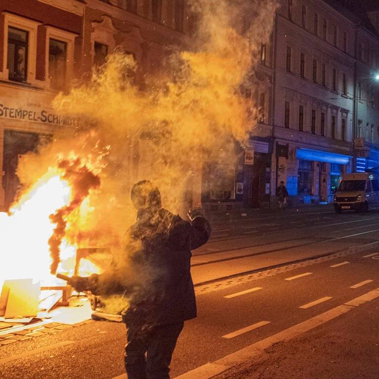 Eine vermummte Person steht neben einer brennenden Barrikade in Leipzig-Connewitz und hält eine Rauchbombe oder einen Molotow-Cocktail in der Hand. (Foto: IMAGO, IMAGO/Moritz Schlenk)