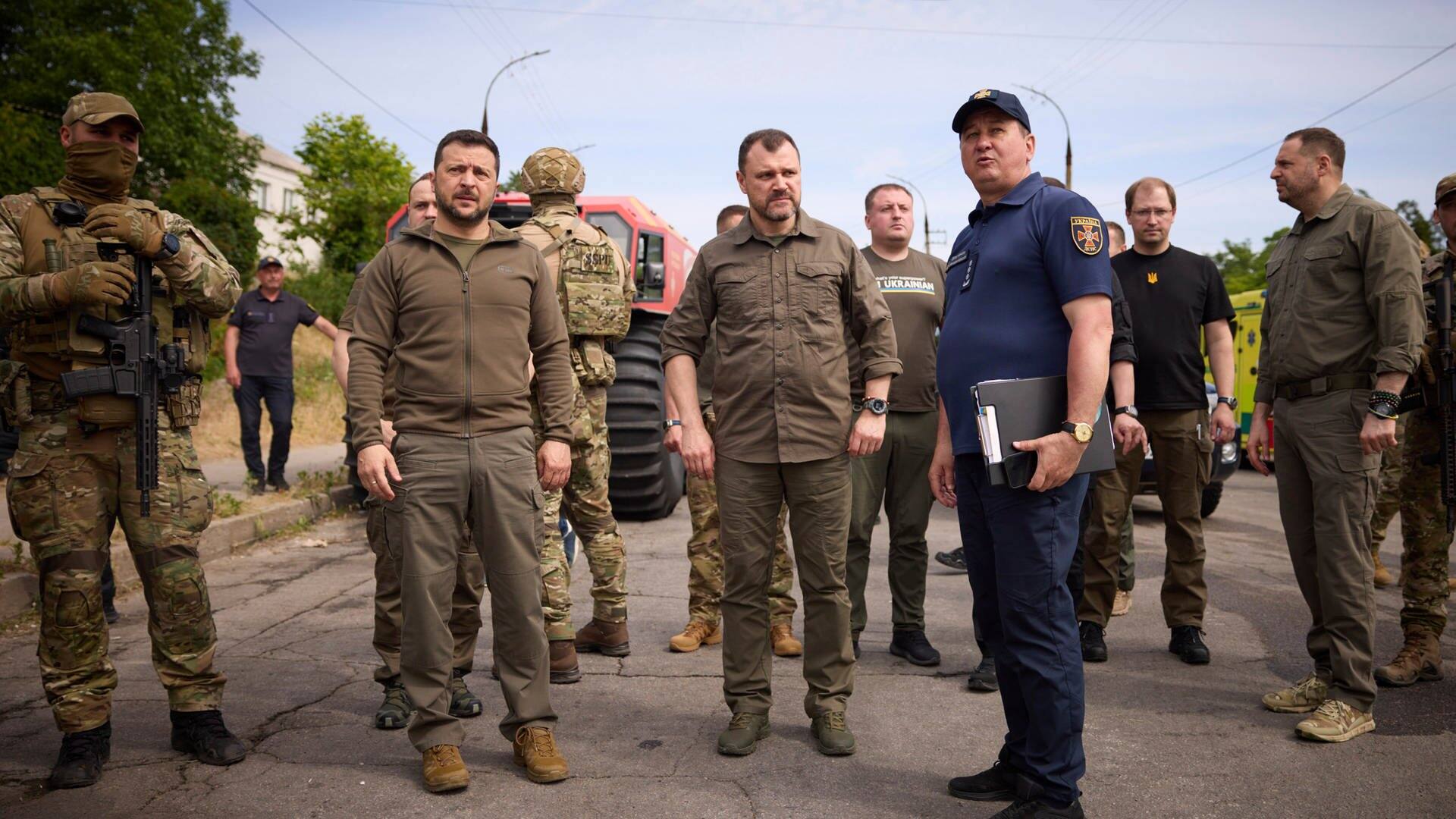 Wolodymyr Selenskyj, Präsident der Ukraine, steht mit anderen Regierungsvertretern auf einer Straße und besucht die von den Überschwemmungen betroffenen Gebiete in Cherson. (Foto: dpa Bildfunk, picture alliance/dpa/Ukrainian Presidential Press Office/AP | Uncredited)