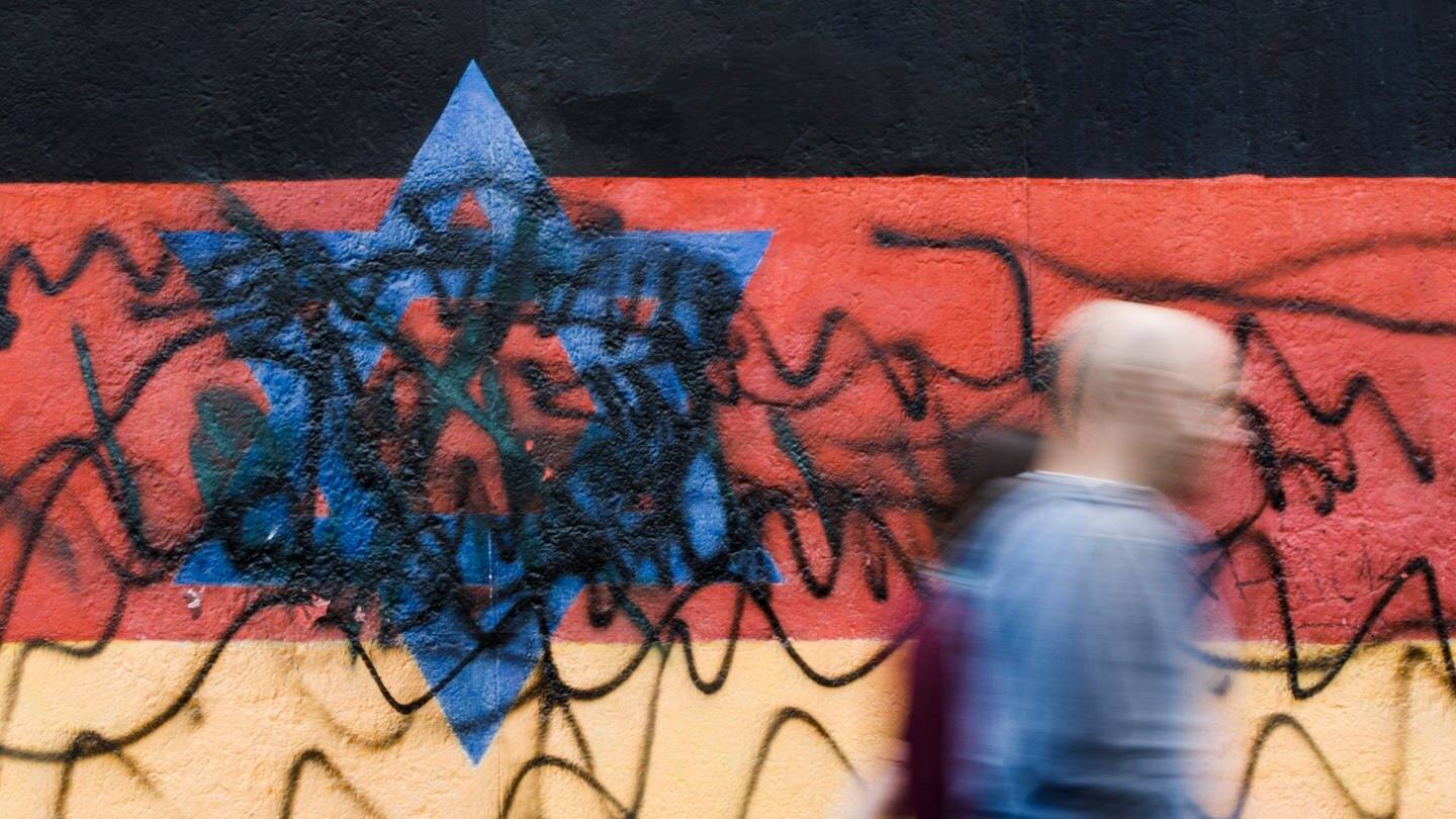 Berlin: Passanten gehen an dem zerstörten Wandbild des Künstlers Günther Schäfer an der East Side Gallery vorbei. Das Wandbild „Vaterland“, das die schwarz-rot-goldene Deutschlandfahne mit einem blauem Davidstern zeigt, wurde von Unbekannten mit schwarzen Graffitis beschmiert. (Foto: dpa Bildfunk, picture alliance/dpa | Carsten Koall)