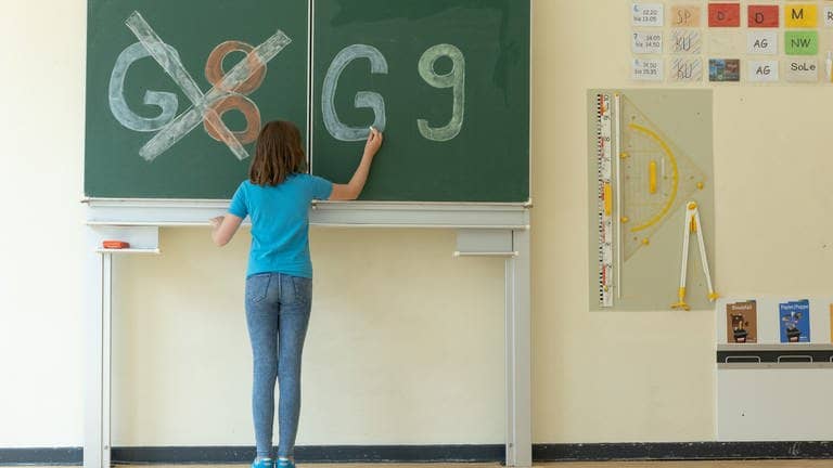 Ein Mädchen schreibt an eine Tafel. G8 ist durchgestrichen, auf der rechten Tafelhälfte steht G9. (Foto: picture-alliance / Reportdienste, Picture Alliance)