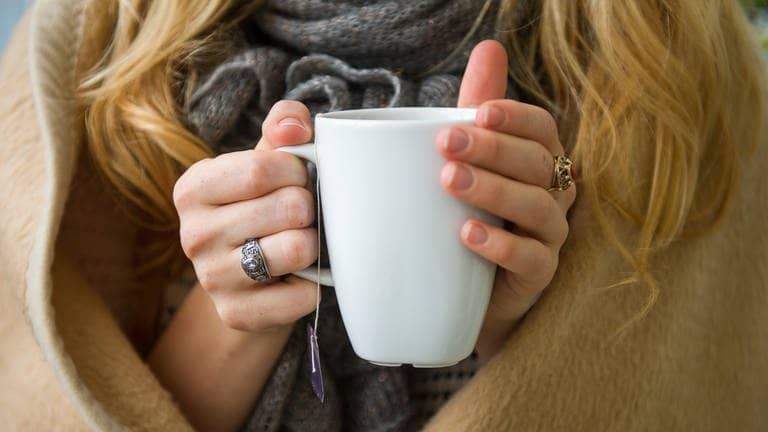 Eine Frau wärmt sich in eine Decke gehüllt, an einer Tasse Tee in den Händen