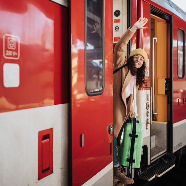 Das 49-Euro-Ticket startet jetzt. Symbolbild: Eine Frau winkt an der Tür eines Zug stehend mit grünem Koffer in der Hand