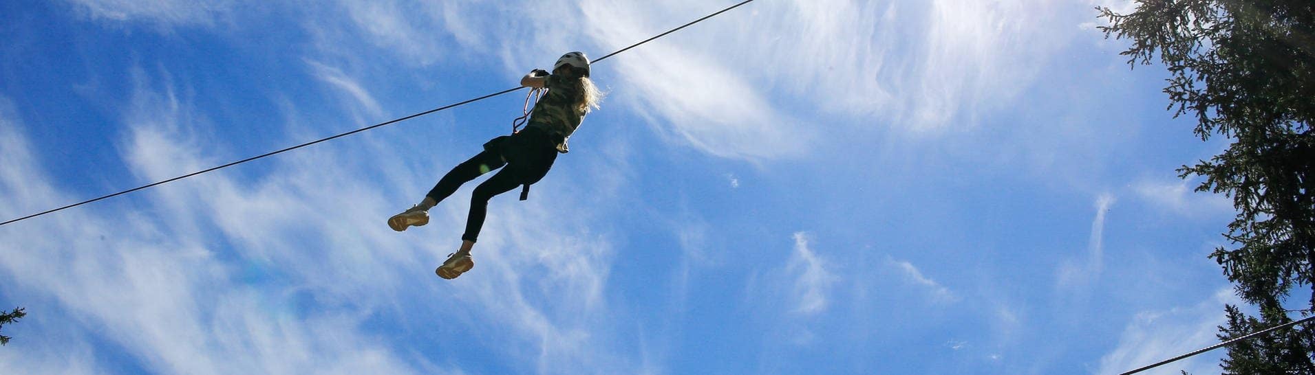 Eine junge Frau gleitet mit einer Seilrutsche vor blauem Himmel (Foto: IMAGO, imago images/Geisser)