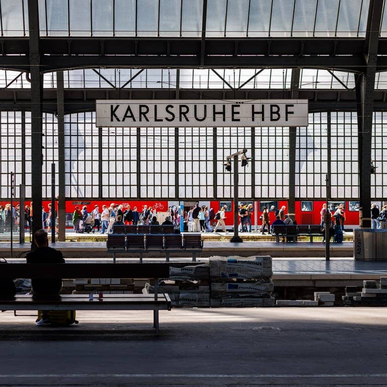 Stromschlag im Zug: Menschen gehen neben einem RE Regionalexpress der DB Deutschen Bahn im Karlsruher Hauptbahnhof über einen Bahnsteig während im Vordergrund Menschen auf einer Bank sitzen.
