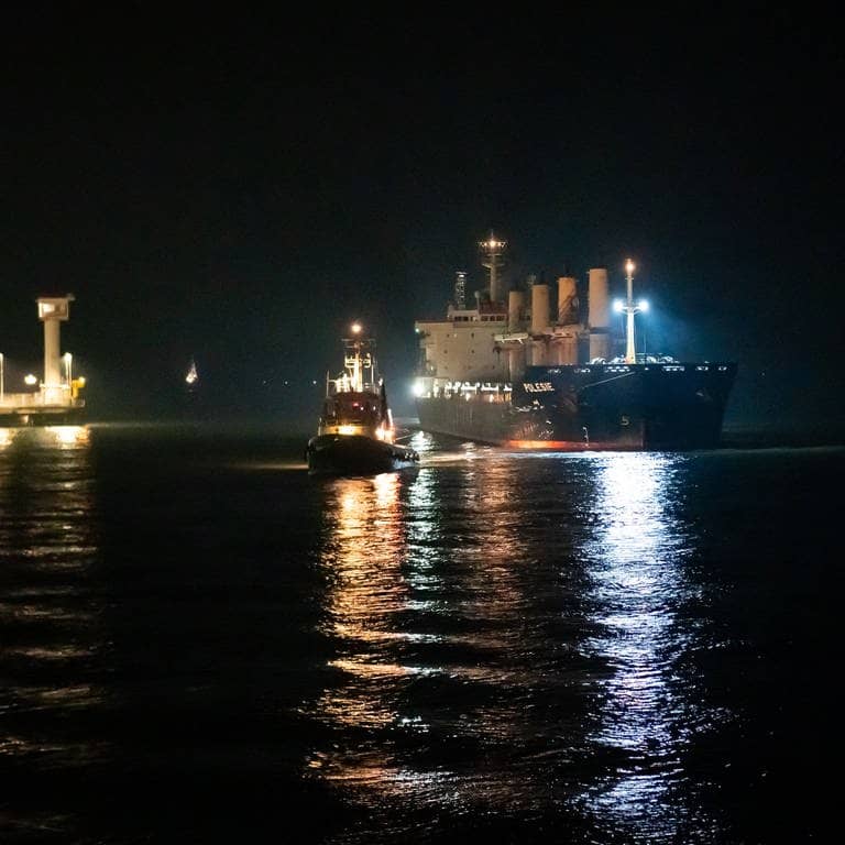 Zwei Schiffe liegen vor einem beleuchteten Hafen. Das Frachtschiff „Polesie“ wird in der Nacht von zwei Schleppern an den Kai der Seebäderbrücke in Cuxhaven gezogen.