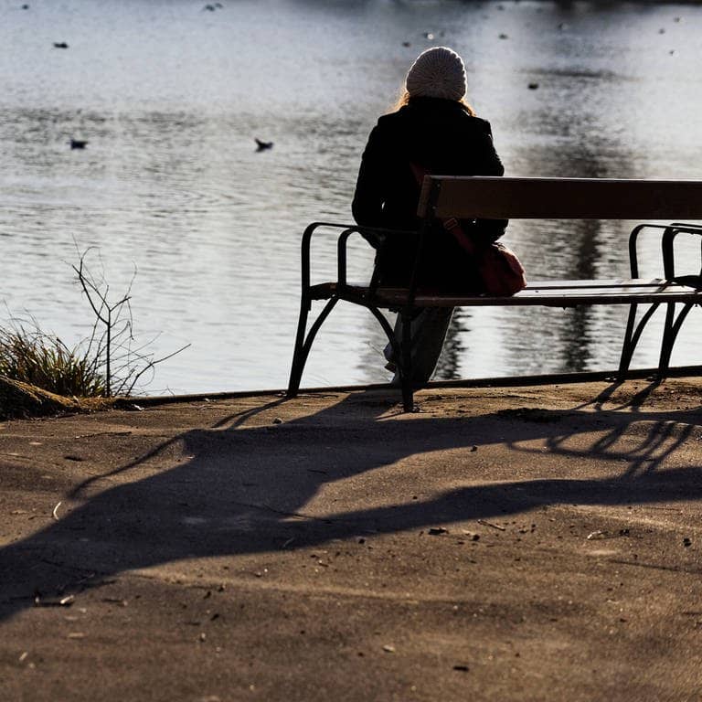 Eine Frau sitzt alleine auf einer Bank an einem See (Foto: IMAGO, imago images/robertkalb photographien)