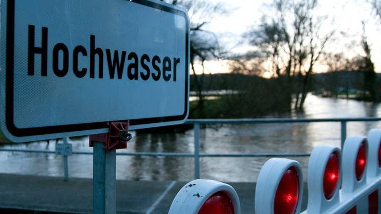 Ein Schild und eine Absperrung weisen auf Hochwasser an einem Fluss hin