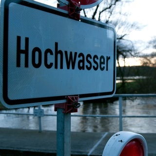Ein Schild und eine Absperrung weisen auf Hochwasser an einem Fluss hin