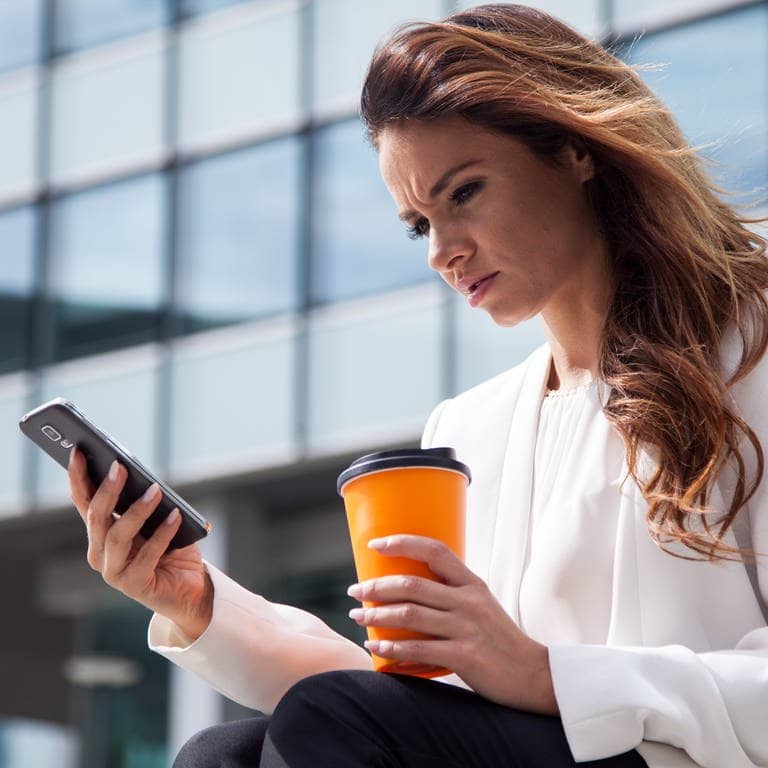 Eine junge Frau mit einem Kaffee in der Hand sitzt am Buerogebaeude auf einer Treppe und schaut irritiert auf ihr Smartphone. (Foto: dpa Bildfunk, dpa Themendienst | Christin Klose)