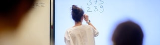 Eine Lehrerin schreibt eine Mathematikaufgabe auf eine digitale Schultafel im Klassenraum einer 4. Klasse einer Grundschule.