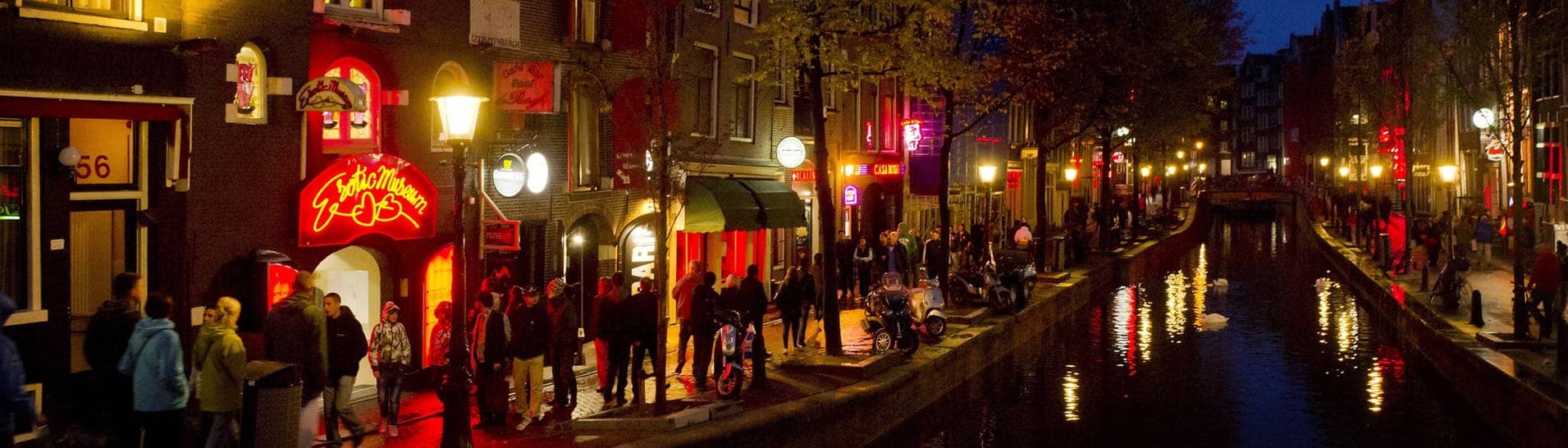 Menschen schlendern in Amsterdam am Abend an einer Gracht entlang durch den Rotlichtbezirk De Wallen.