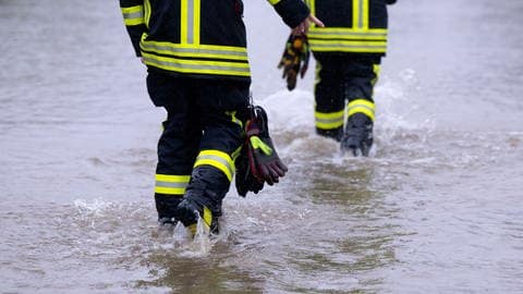 Feuerwehrleute gehen über eine überflutete Straße in Dasing im schwäbischen Landkreis Aichach-Friedberg.