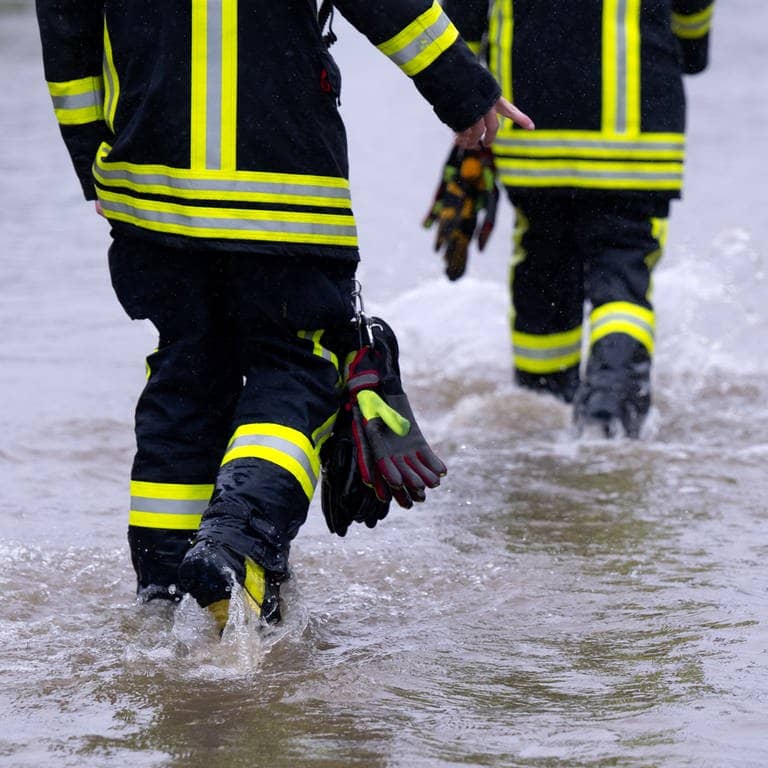 Feuerwehrleute gehen über eine überflutete Straße in Dasing im schwäbischen Landkreis Aichach-Friedberg.
