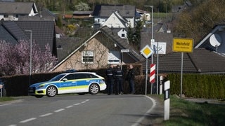 Ein Polizeiauto und Polizisten stehen am Ortseingang vor Weitefeld. Dort gab es einen mutmaßlichen Dreifachmord, der mutmaßliche Täter ist noch auf der Flucht.
