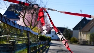 Mit Flatterband der Polizei ist eine Straße abgesperrt. Die Ermittlungen zu der Gewalttat mit drei toten Familienmitgliedern im Westerwald gehen weiter.