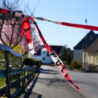 Mit Flatterband der Polizei ist eine Straße abgesperrt. Die Ermittlungen zu der Gewalttat mit drei toten Familienmitgliedern im Westerwald gehen weiter.