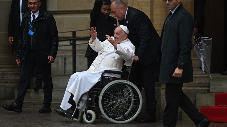 Papst Franziskus wird im Rollstuhl geschoben und streckt die Arme in die Höhe.