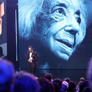 Pianist Igor Levit steht auf der Bühne des Deutschen Filmpreises und hält eine Trauerrede für die gestorbene Margot Friedländer, von der Foto im Hintergrund auf einer Leinwand zu sehen ist.
