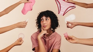 Eine Frau schaut nachdenklich zur Seite, viele Hände halten ihr Produkte für die Periode hin. Menstruation ist noch immer ein Tabuthema.