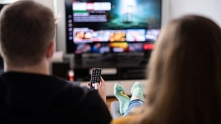 Symbolbild: Klage gegen Netflix wegen Preiserhöhung. Man sieht auf dem Bild ein Paar, einen Nann und eine Frau von hinten, die Fernsehen schauen.
