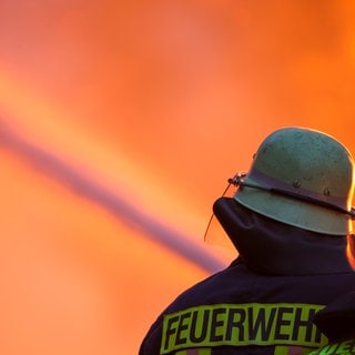Symbolbild Feuerwehr beim Löschen. Man sieht einen Feuerwehrmann mit Schlauch und im Hintergrund ein großes Feuer.