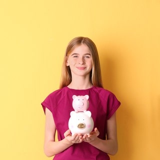 Symboldbild eines Mädchens mit einem Sparschwein in der Hand, das für Kinder steht, die Taschengeld auf eine Bezahlkarte, Kreditkarte oder in bar erhalten.