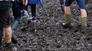 Menschen mit Gummistiefeln laufen durch Schlamm. Das Wetter während dem Rock am Ring Festival könnte verregnet werden.