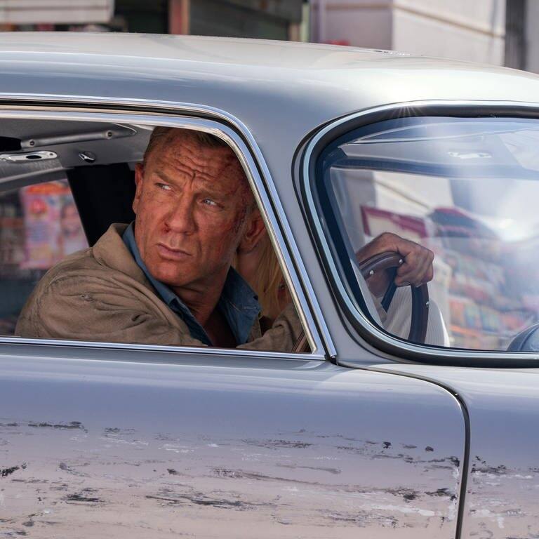 Schauspieler Daniel Craig sitzt als Agent James Bond an einem Filmset in einem demolierten Auto. (Foto: dpa Bildfunk, picture alliance/dpa/EON Productions/Metro-Goldwyn-Mayer Studios | Nicola Dove)