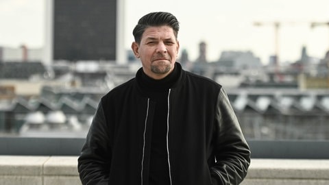 Tim Mälzer in einer Lederjacke auf einer Terasse vor Großstadtkulisse