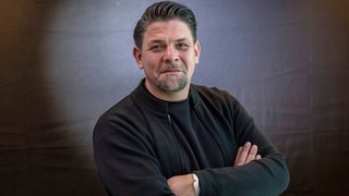 Tim Mälzer blickt mit verschränkten Armen frontal in die Kamera