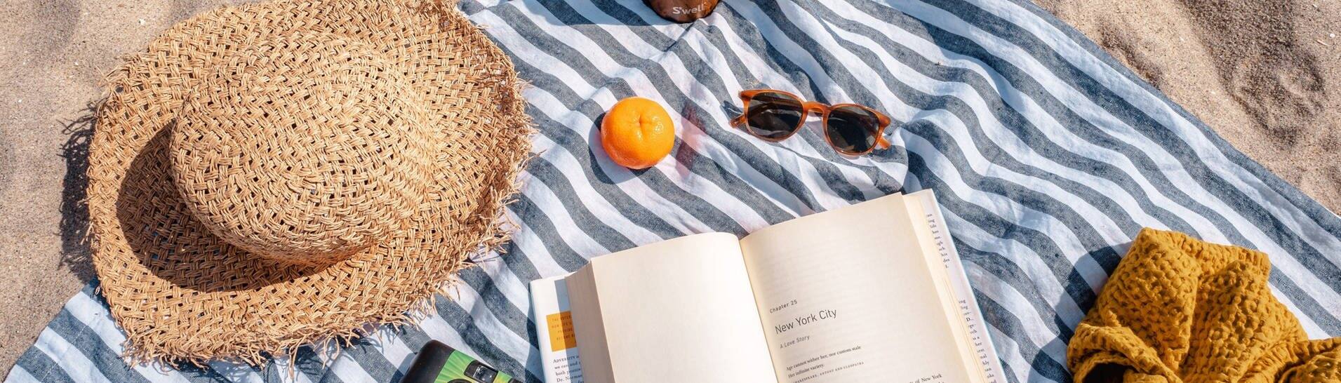 Sonnenhut, Sonnenbrille und Buch auf einer Decke am Meer (Foto: Photo by S'well on Unsplash)