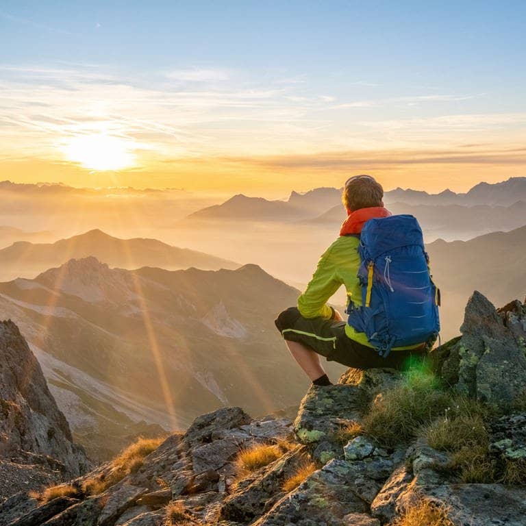 Junger Wanderer sitzt mit Rucksack auf einem Stein und blickt in das Berggipfel-Panorama (Foto: Adobe Stock/Netzer Johannes)