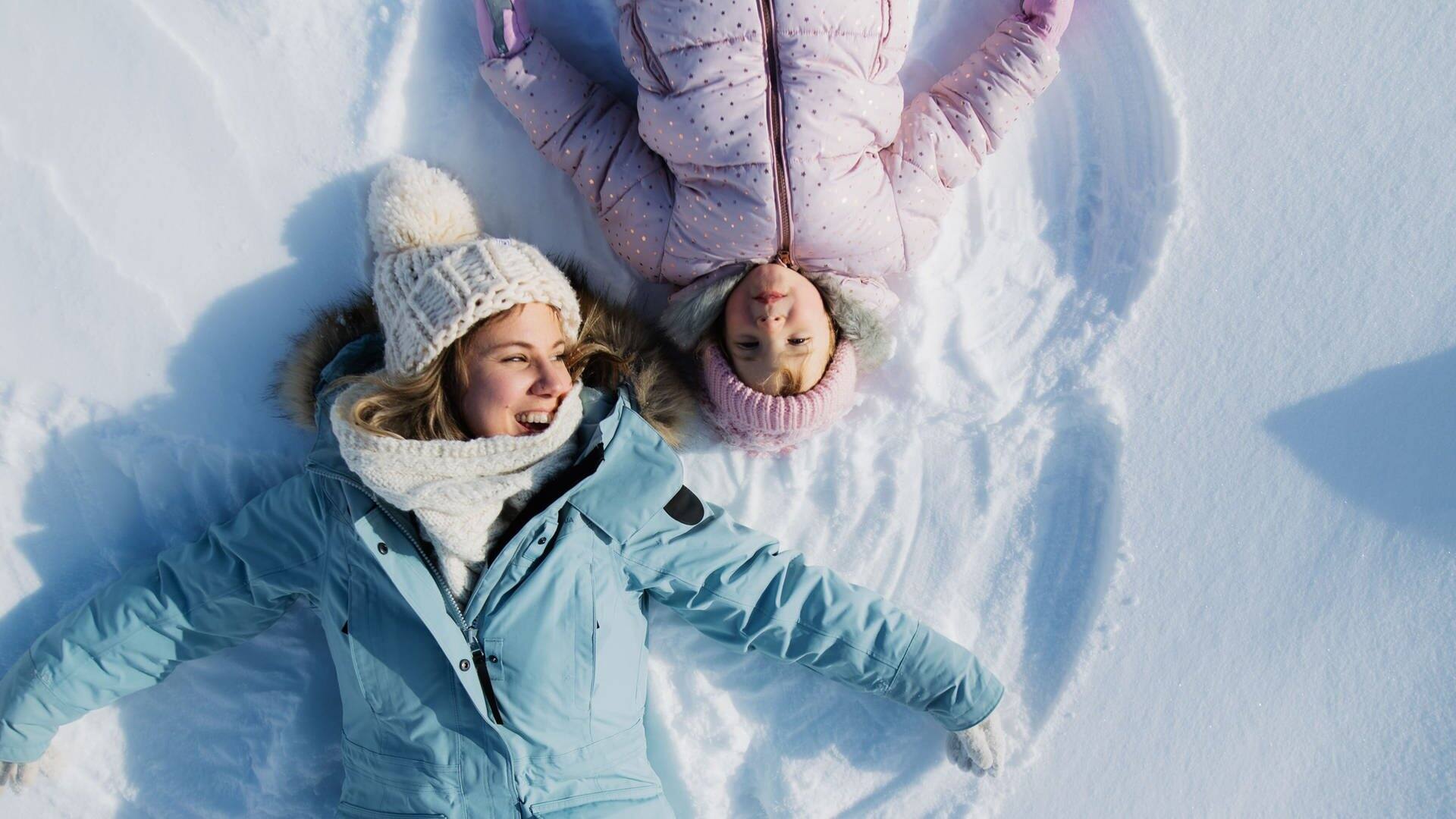Mutter und Kind liegen im Schnee, lachen und machen einen Schneeengel (Foto: Adobe Stock/Halfpoint)