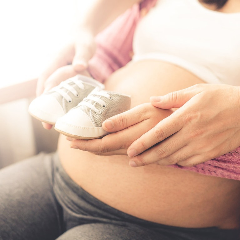 Eine Frau mit Babybauch, die zusammen mit ihrem Partner kleine Babyschühchen darauf hält. (Foto: IMAGO, IMAGO / ingimage)