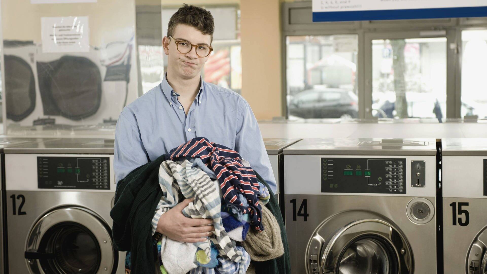 Mann schaut komisch aus der Wäsche, mit Wäsche in der Hand in einem Waschsalon (Foto: IMAGO, Imago)