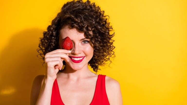 Junge Frau mit braunen Locken hält sich eine rote Erdbeere vors Gesicht und lacht (Foto: Adobe Stock, deagreez)
