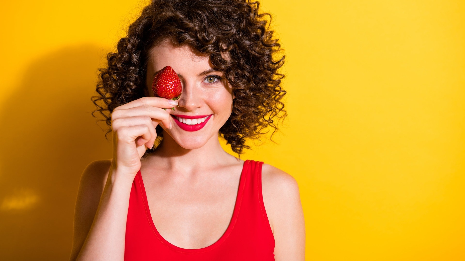 Junge Frau mit braunen Locken hält sich eine rote Erdbeere vors Gesicht und lacht (Foto: Adobe Stock, deagreez)