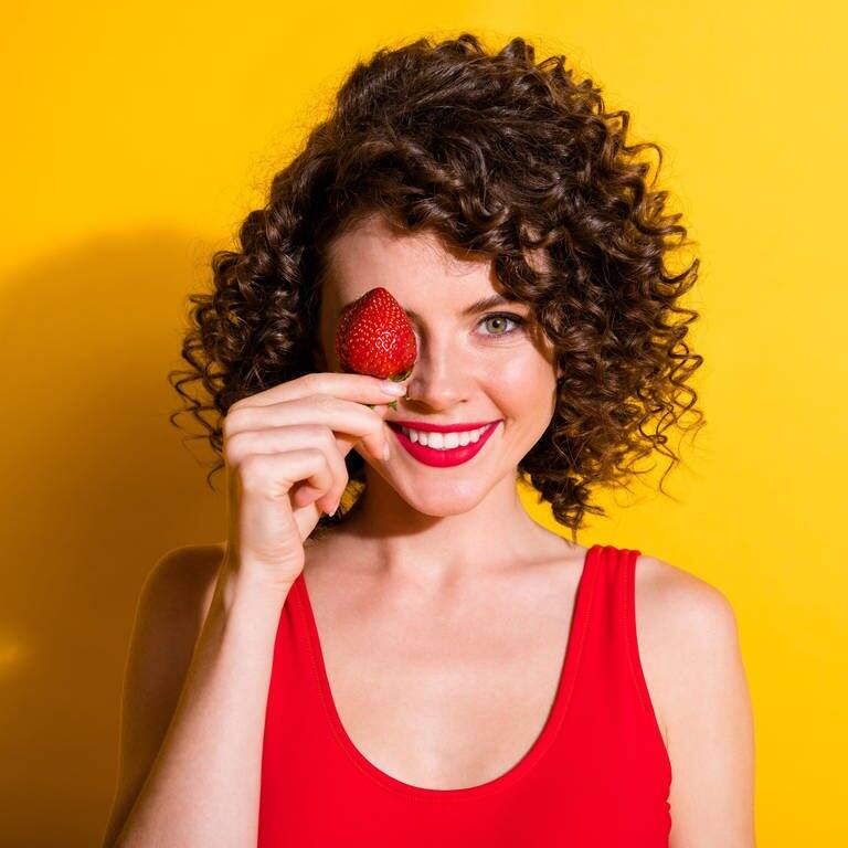 Junge Frau mit braunen Locken hält sich eine rote Erdbeere vors Gesicht und lacht (Foto: Adobe Stock, deagreez)