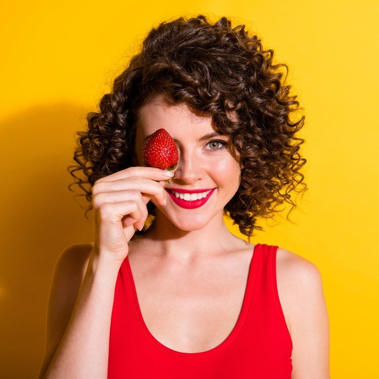 Junge Frau mit braunen Locken hält sich eine rote Erdbeere vors Gesicht und lacht (Foto: Adobe Stock, deagreez)