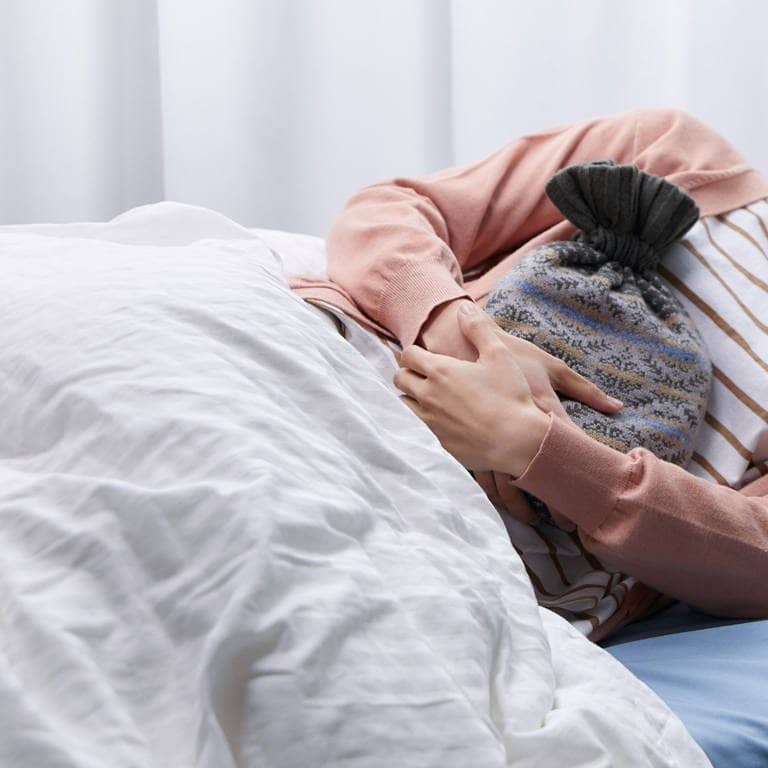 Eine Frau liegt im Bett und hält eine Wärmflasche an ihren Bauch, um ihre Regelschmerzen zu lindern (Foto: Imago)